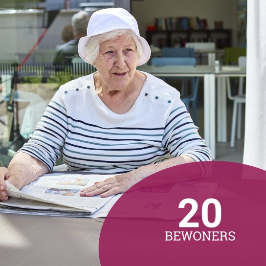 Oudere vrouw met witte hoed en gestreept shirt leest krant aan tafel in lichte ruimte. (vergroot foto)